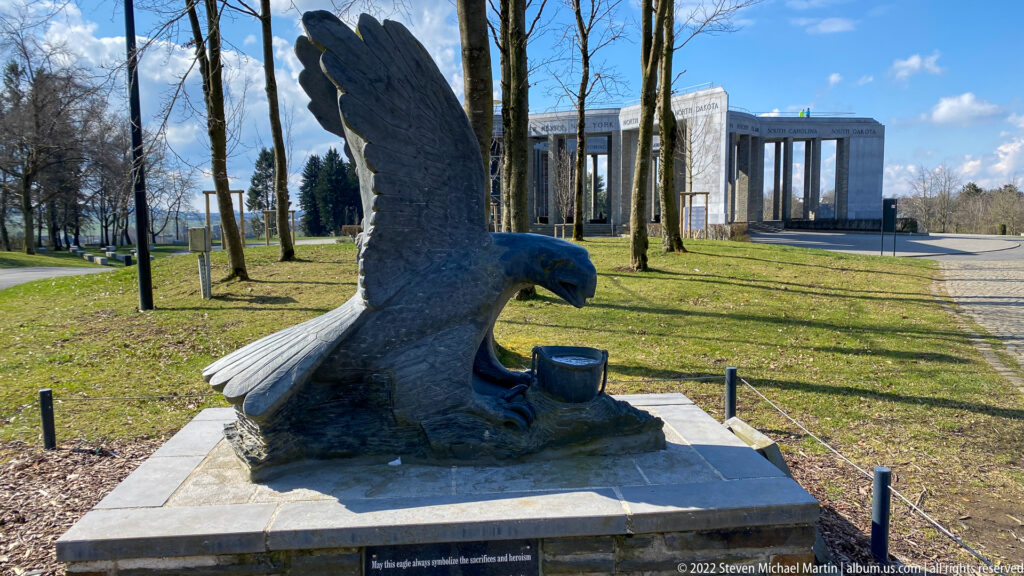 Large eagle sculpture with raised wings on a stone base, with the Mardasson Memorial visible in the background.