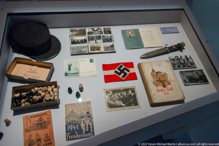 Display case with photographs, documents, cap, small objects, a swastika armband, booklets, and a sheathed tool.