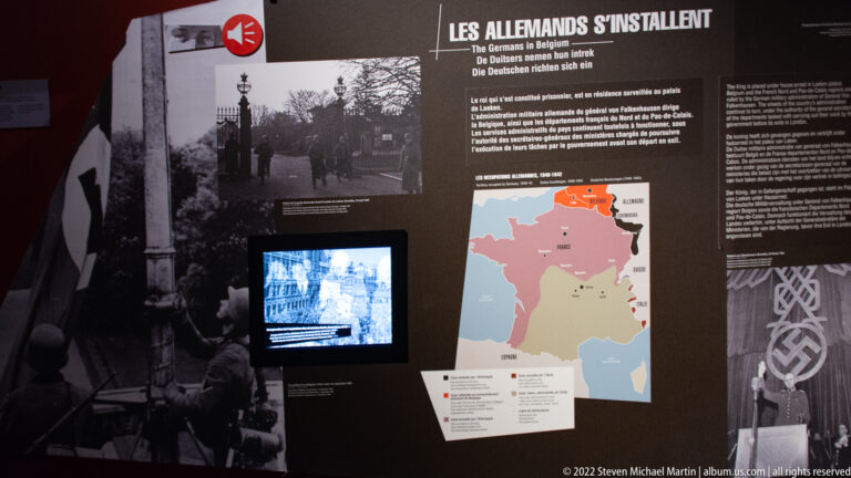 Dark museum panel on the German occupation of Belgium with text, map, photographs, swastika imagery, and video screen.