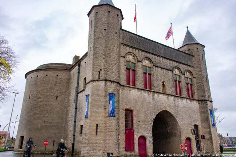 Historic Bruges gatehouse with arched passage, flanking towers, red shutters, flags, and pale brick masonry under a cloudy sky.