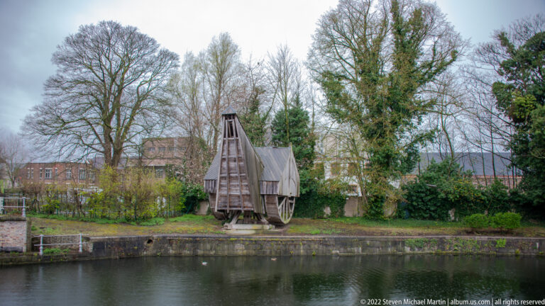 Small timber wheel mechanism beside a canal or basin, framed by trees and masonry banks in Bruges.