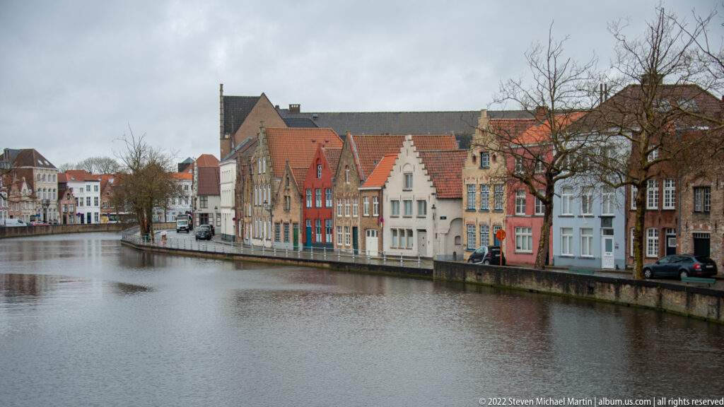 Broad Bruges canal with rows of colorful gabled houses, bare trees, and parked cars along the quay.