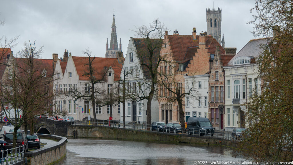 Canal-front houses in Bruges with a slender church spire and a broad square tower rising behind the roofs.