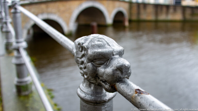 Close view of a sculpted metal animal-head finial on a white canal railing with water blurred behind.