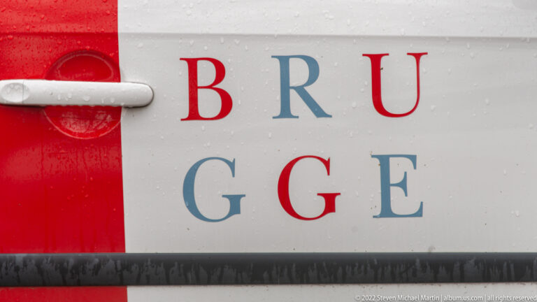 Close-up of red and blue letters spelling BRUGGE on a rain-speckled white vehicle or panel.