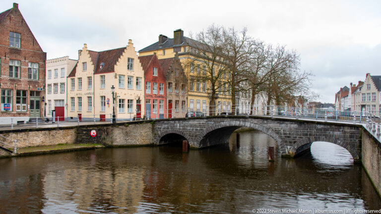 Three-arch stone bridge over a Bruges canal with stepped-gable houses and bare trees beyond.