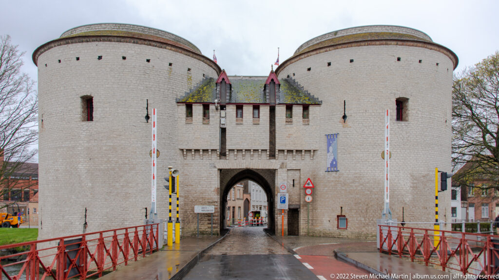 Twin round towers flank a central arched gateway at the end of a bridge with red railings in Bruges.