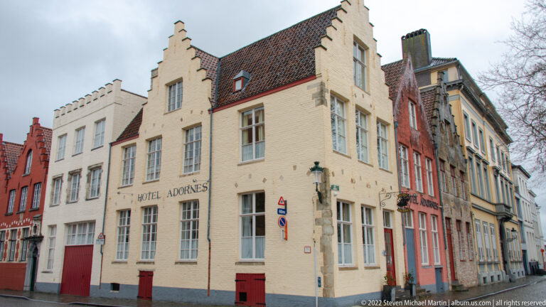 Pale stepped-gable façade of Hotel Adornes on a Bruges street corner with red doors and tall windows.