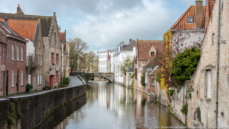 Narrow Bruges canal between brick houses and garden walls, with a small bridge in the distance.