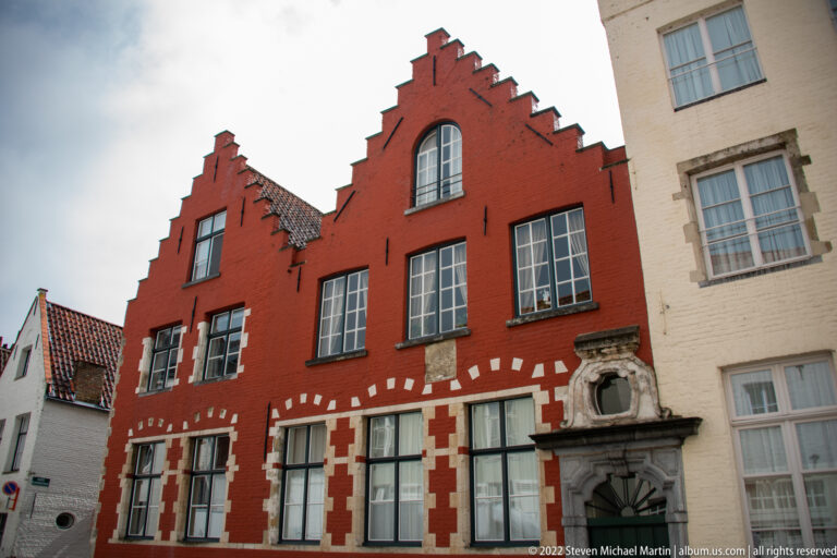 Red stepped-gable townhouse with white stone trim, large windows, and carved doorway surround in Bruges.