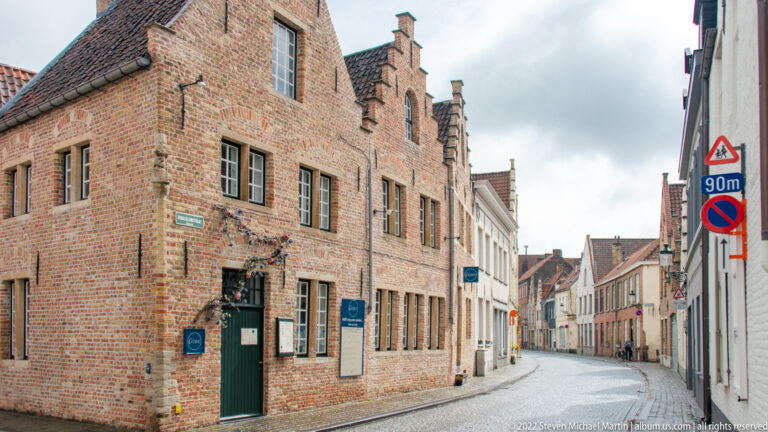 Large stepped-gable brick building beside a narrow quiet lane in Bruges, with pale houses receding in the distance.