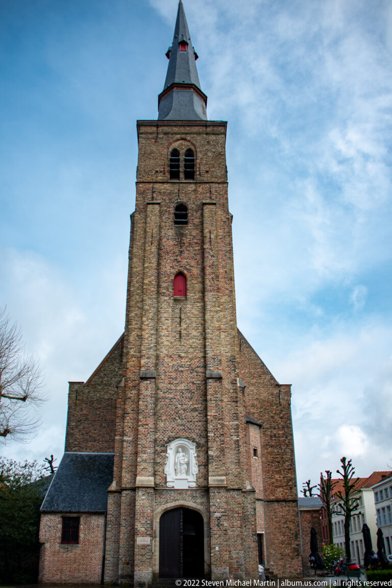 Brick church with square tower, pyramidal roof, paired belfry openings, and arched entrance in Bruges.