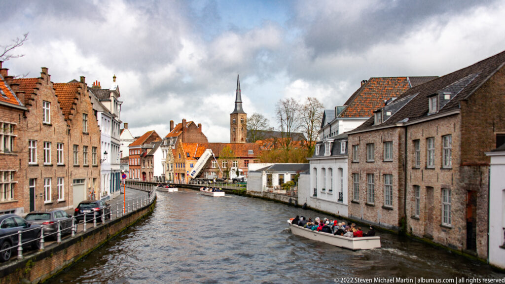 Passenger boat moves along a narrow Bruges canal between historic houses, with a church tower in the distance.
