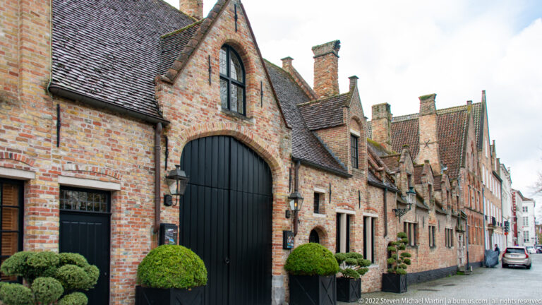 Historic brick houses line a narrow Bruges lane, led by a tall black arched carriage doorway and clipped shrubs.