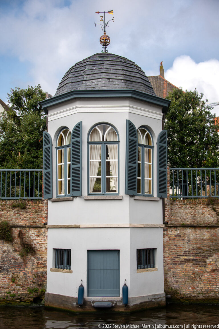 White octagonal pavilion with arched windows, shutters, slate roof, and ship weather vane against a brick wall in Bruges.