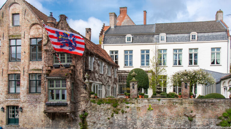 Weathered brick courtyard building with heraldic flag facing a pale townhouse and clipped trees in Bruges.