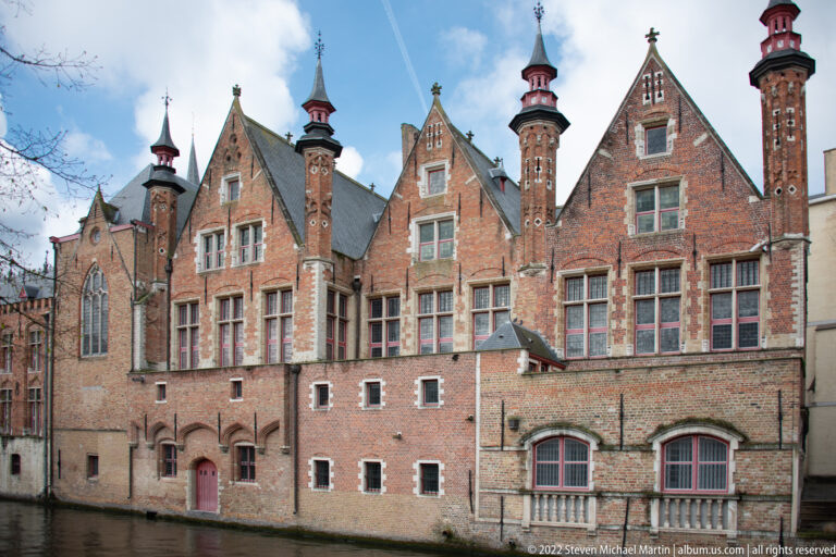 Brick civic buildings with steep gables, tall windows, and multiple ornate chimney stacks in Bruges.