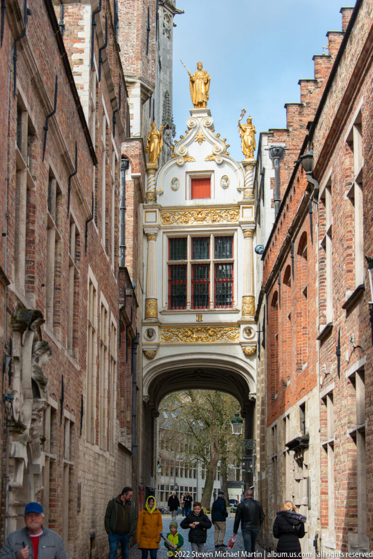 White-and-gold decorated arched passage bridges a narrow Bruges street, with pedestrians walking beneath.