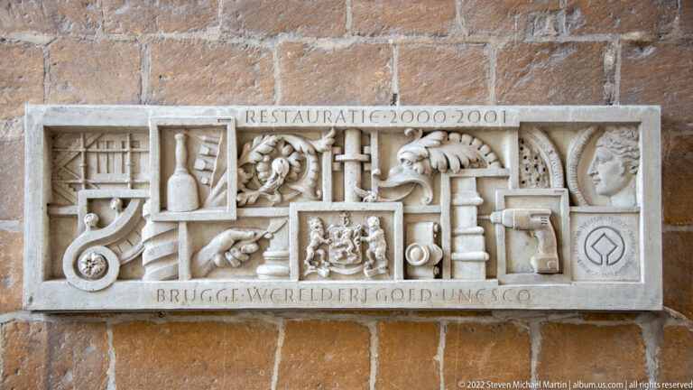 Carved stone relief dated 2000–2001 with restoration motifs and Brugge Werelderfgoed UNESCO inscription on brick wall.