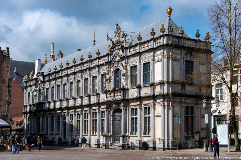 Grand pale-stone historic building with tall windows, balustrade, and rooftop sculptures on a Bruges square.