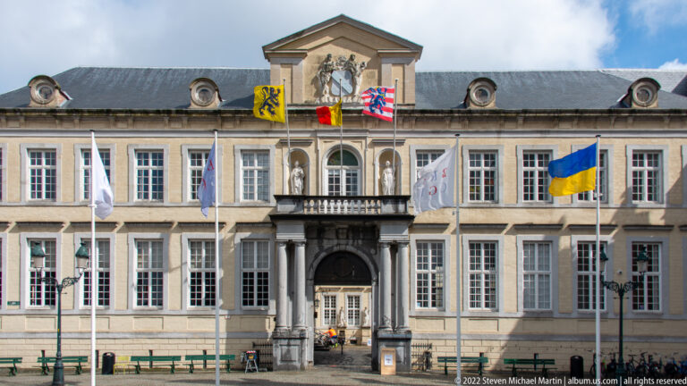 Symmetrical neoclassical building with central pediment, columns, heraldic shields, and several flags in Bruges.