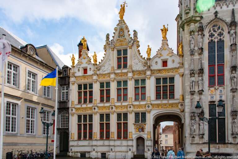 Ornate white-and-gold gabled building with arched passage beside a tall gray Gothic civic façade in Bruges.