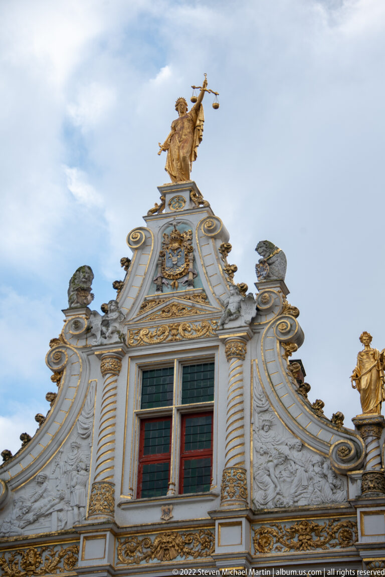Close view of white sculpted gable with gold scrollwork, twisted columns, red windows, and crowning figure in Bruges.