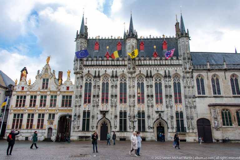 Bruges City Hall with Gothic traceried façade, pinnacles, and flags beside an ornate white-and-gold civic building.