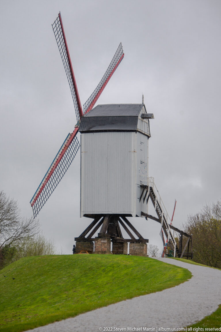 Wooden post mill with lattice sails and side stair standing on a grassy embankment under an overcast sky.
