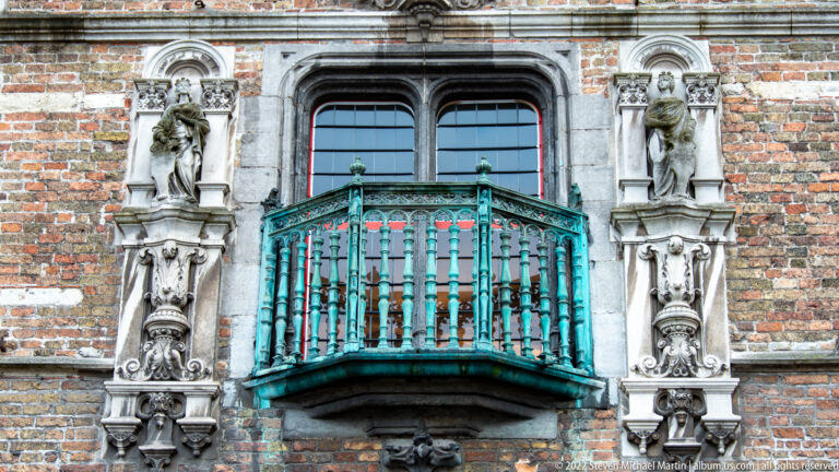Green projecting balcony with red-trimmed windows and carved stone figures on a brick façade in Bruges.