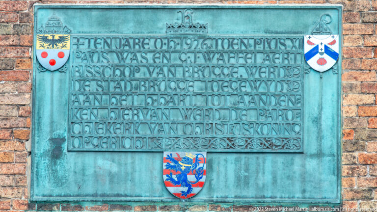 Green-patinated bronze plaque with raised Dutch inscription and heraldic shields mounted on a brick wall in Bruges.