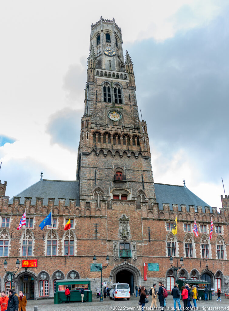 Belfry of Bruges rising above the Markt, with clock faces, brick arcades, flags, and people below.