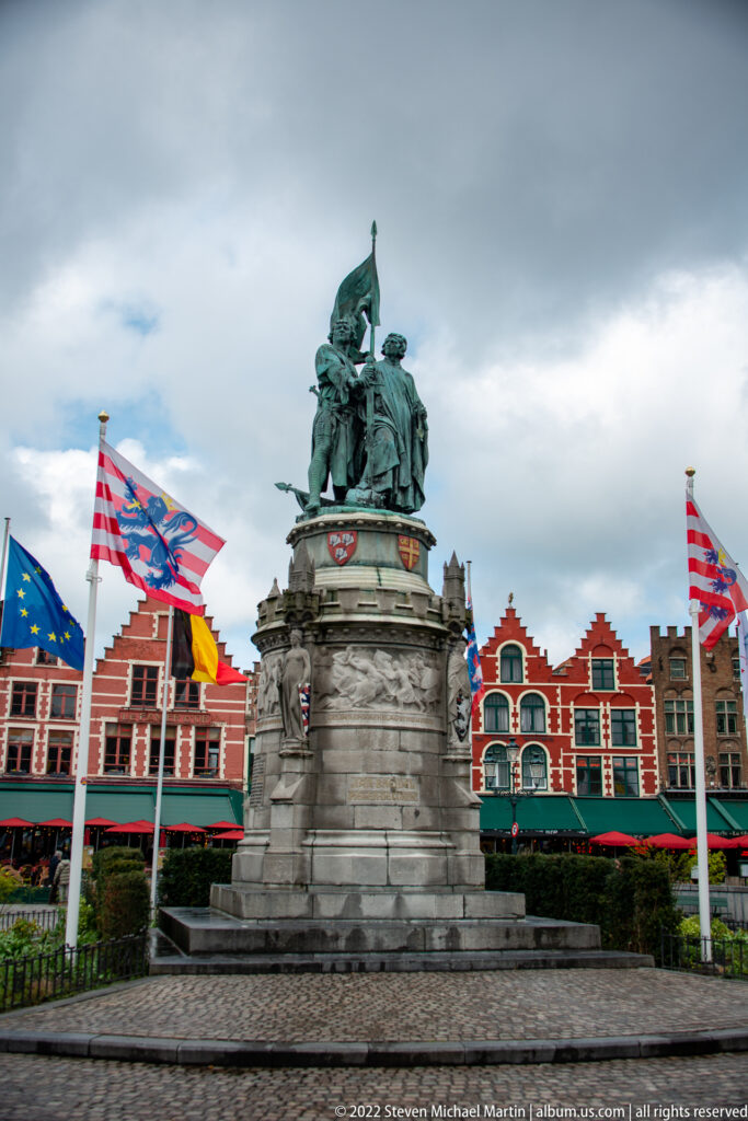 Bronze monument to Jan Breydel and Pieter de Coninck on a stone pedestal amid flags and gabled buildings in Bruges.