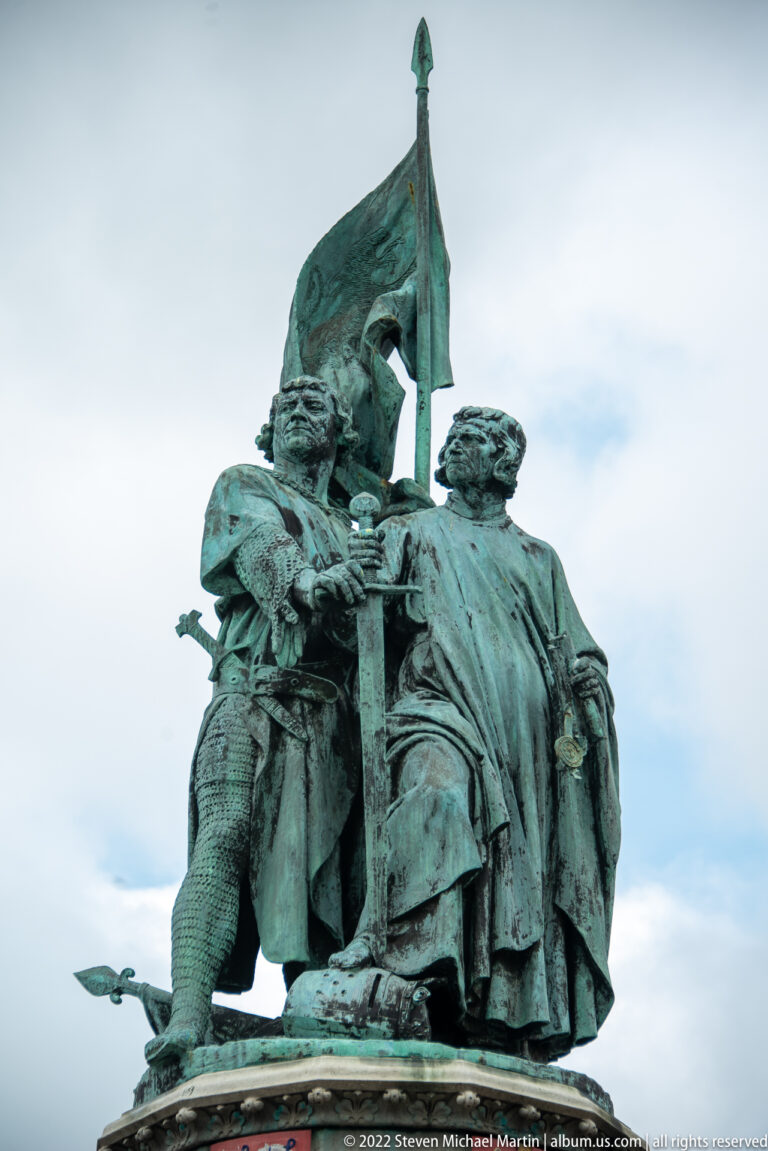Close view of bronze figures of Jan Breydel and Pieter de Coninck in medieval dress beneath a raised banner.