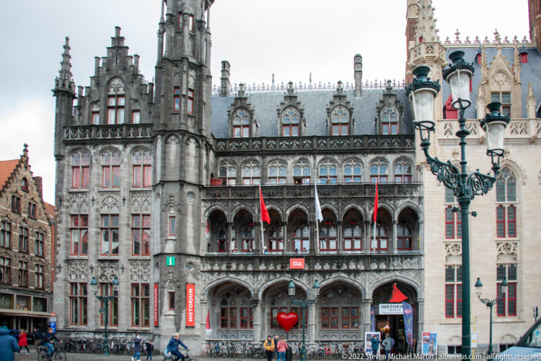 Ornate stone façade of Historium Brugge with Gothic detailing, arcades, red signage, and adjoining civic buildings.
