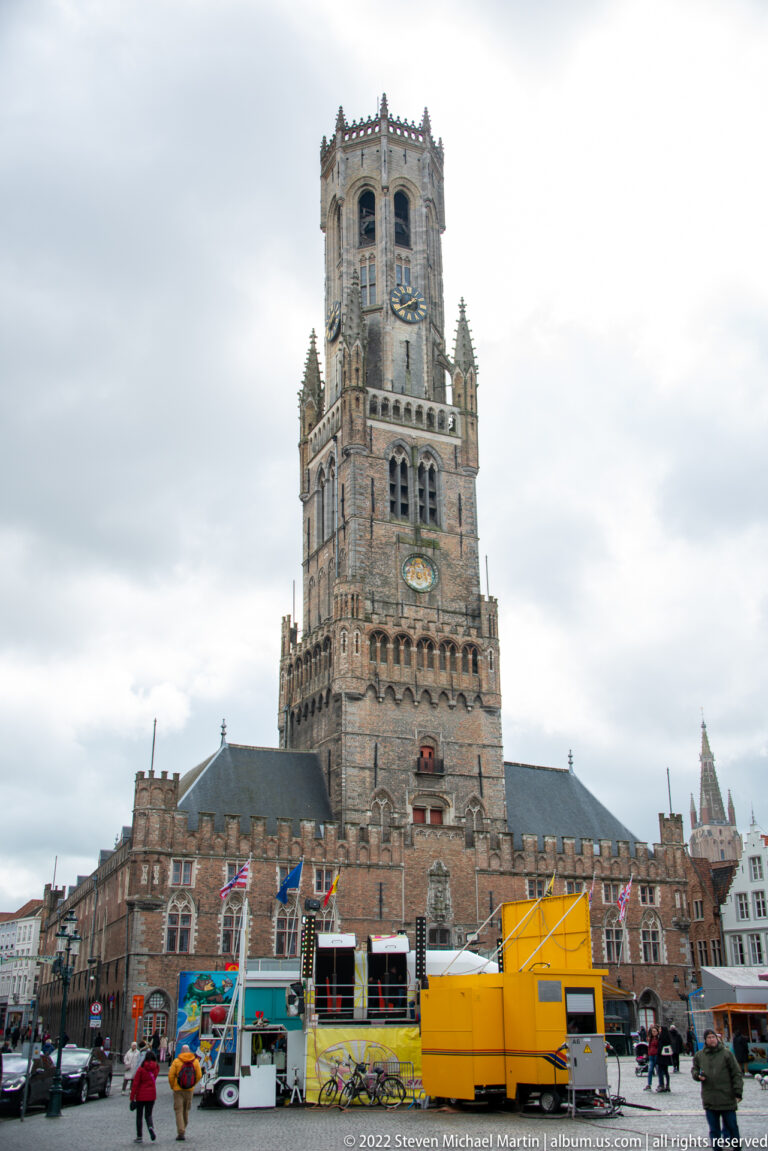 Belfry of Bruges above yellow market vehicles, stalls, flags, and pedestrians on the Markt.