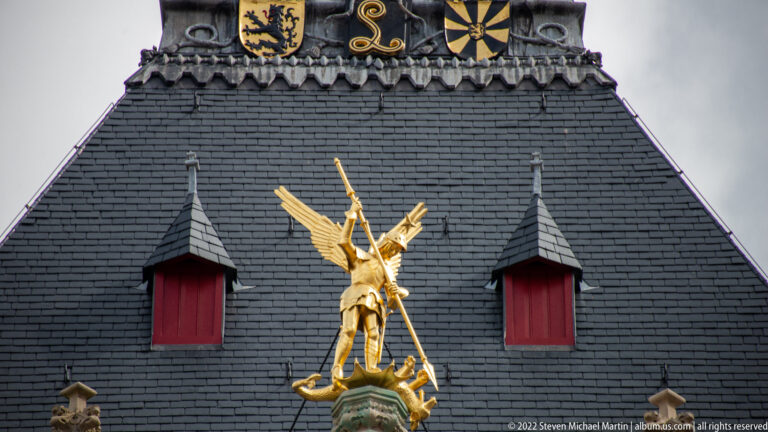 Golden winged figure with staff above a secondary figure, set between red dormers on a dark slate roof in Bruges.