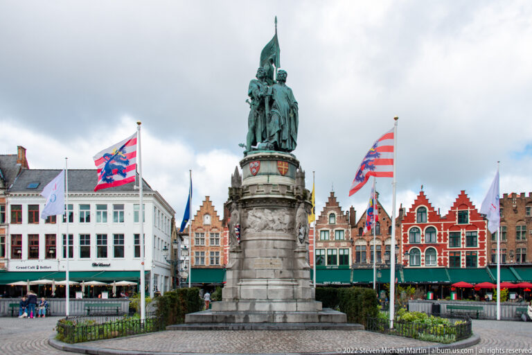 Jan Breydel and Pieter de Coninck monument amid flags and colorful stepped-gable buildings on the Bruges Markt.