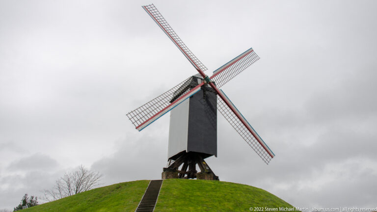 Dark wooden windmill with four lattice sails above a steep grassy mound and central stair in Bruges.
