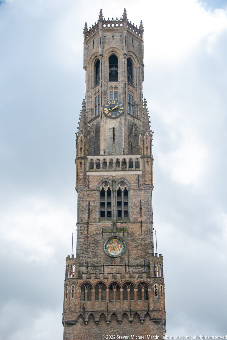Close view of the Belfry of Bruges upper stages with clocks, belfry openings, crenellations, and octagonal crown.