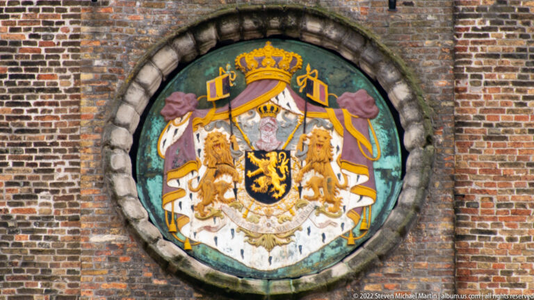 Round painted coat of arms with crowned black-lion shield and supporting lions set into a brick wall in Bruges.
