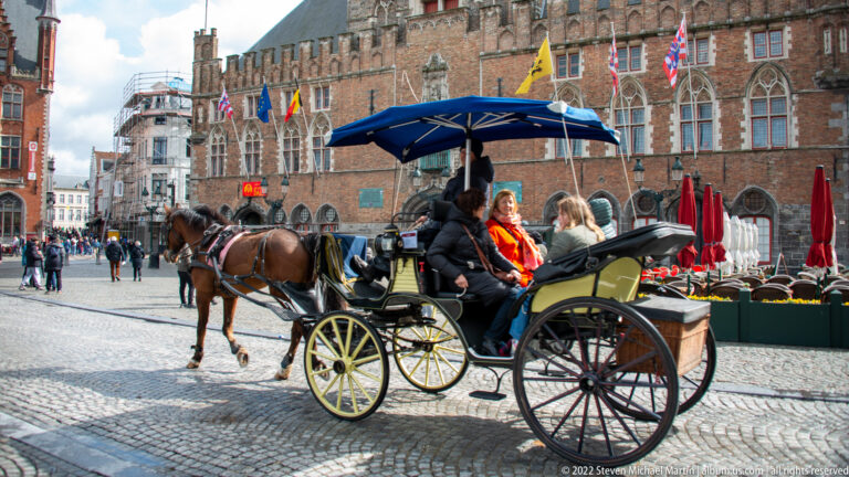 Horse-drawn carriage with passengers crosses the cobbled Bruges Markt before historic brick civic buildings.