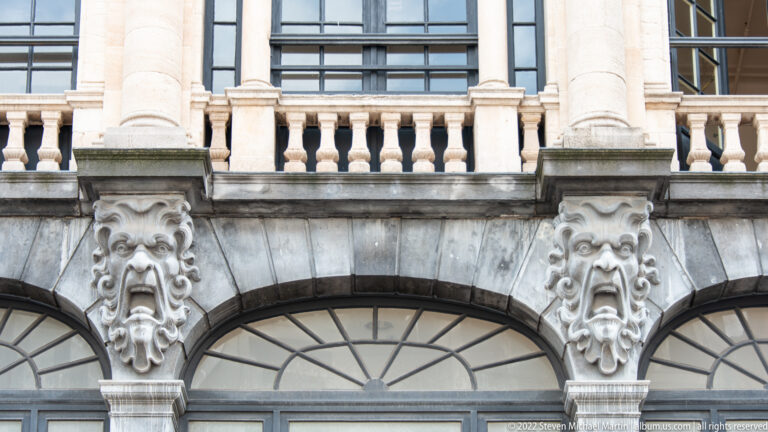 Two carved grotesque masks between arched windows beneath a balustrade on a pale stone Bruges façade.