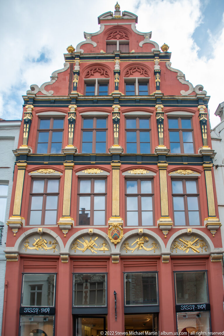 Red-and-gold gabled commercial building with gilded pilasters, tall windows, and ornate upper façade in Bruges.