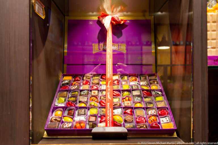 Large open gift box of assorted chocolates arranged in a grid beneath a decorative ribbon in a Bruges shop.