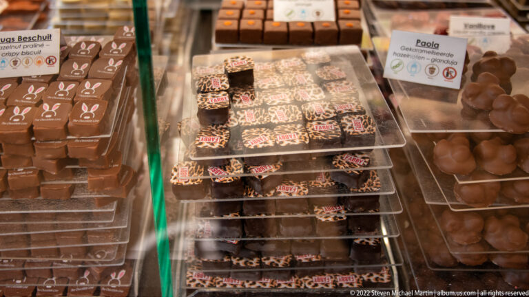 Trays of molded pralines, sprinkle-topped chocolate squares, and rounded chocolates in a Bruges display case.