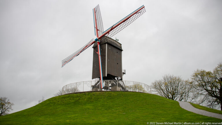 Tall dark wooden windmill with red-edged lattice sails standing on a rounded grassy mound in Bruges.