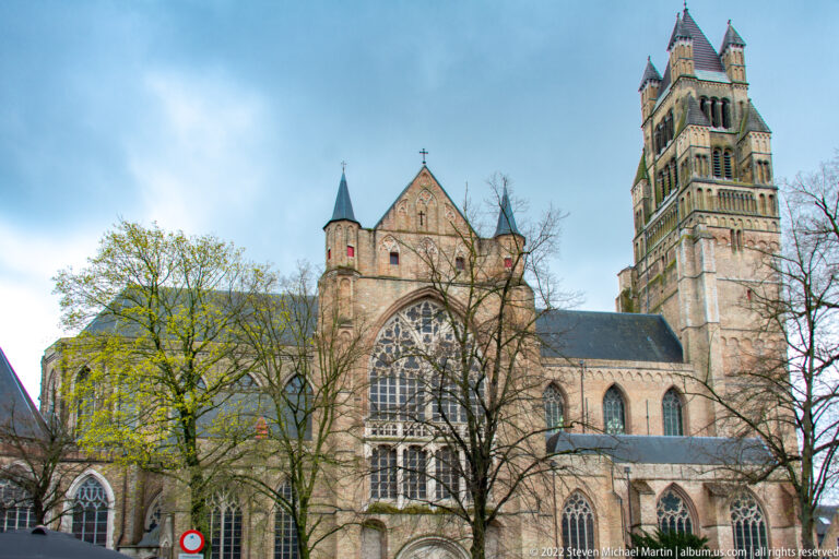 Large Gothic church in Bruges with tall square tower, pointed transept window, buttresses, and trees in front.