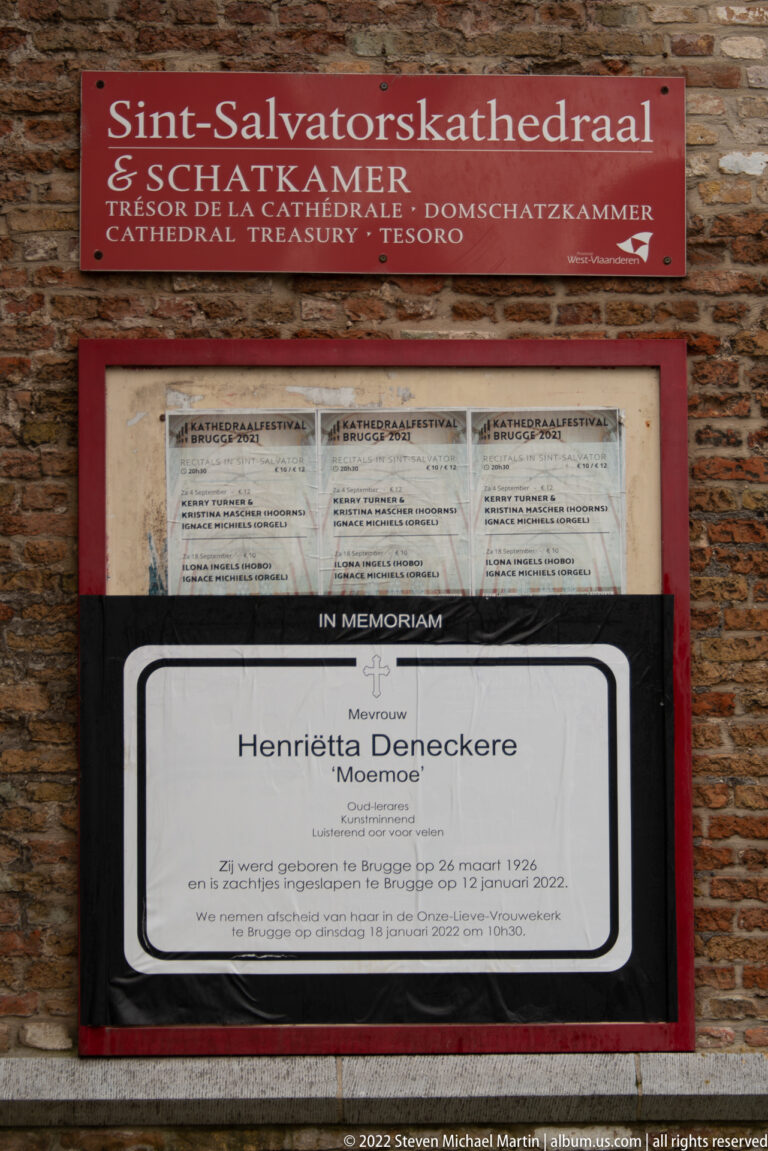 Memorial notice for Henriëtta Deneckere mounted beneath a red Sint-Salvatorskathedraal sign on a brick wall.