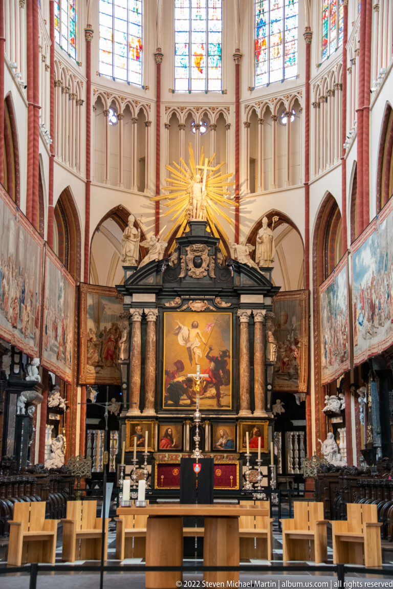 Nave of Saint-Salvator Cathedral with pointed arches, stained glass, paintings, rows of chairs, and ornate high altar.